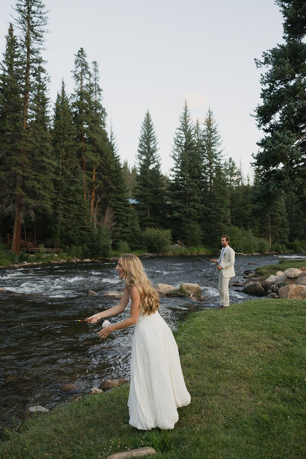 Bride on the bank of the Taylor River with the groom on the rocks behind her