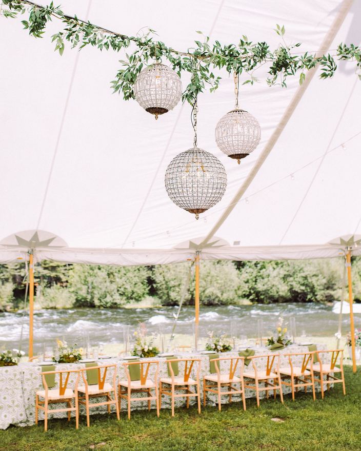 Long reception table under a sailcloth tent with crystal globe chandeliers beside the Taylor River