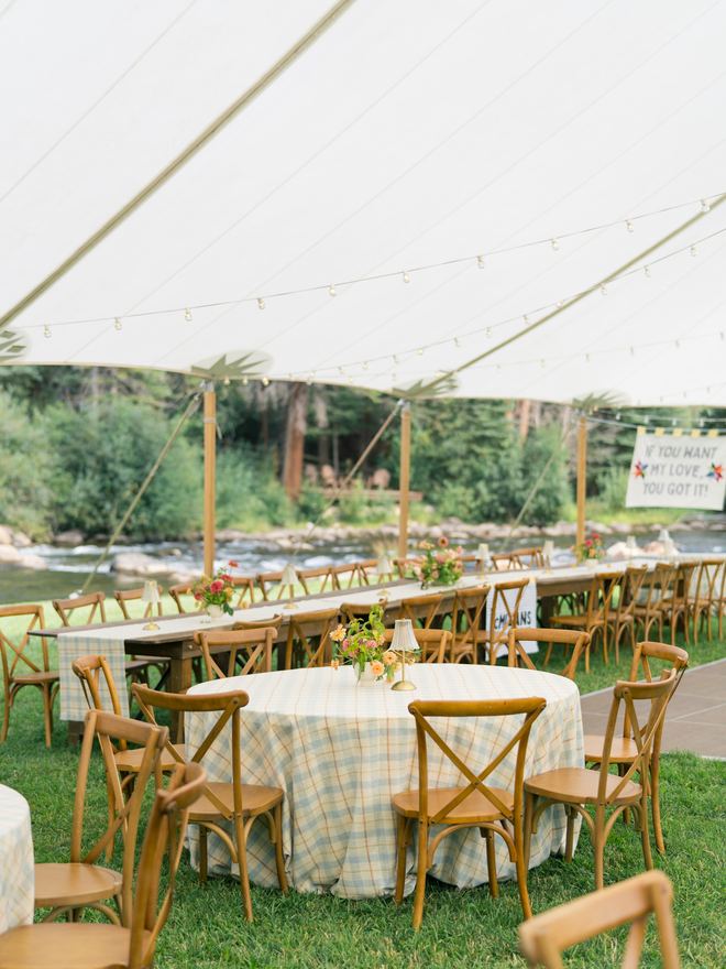 Reception tables with gingham linens and cross-back chairs under a tent by the river