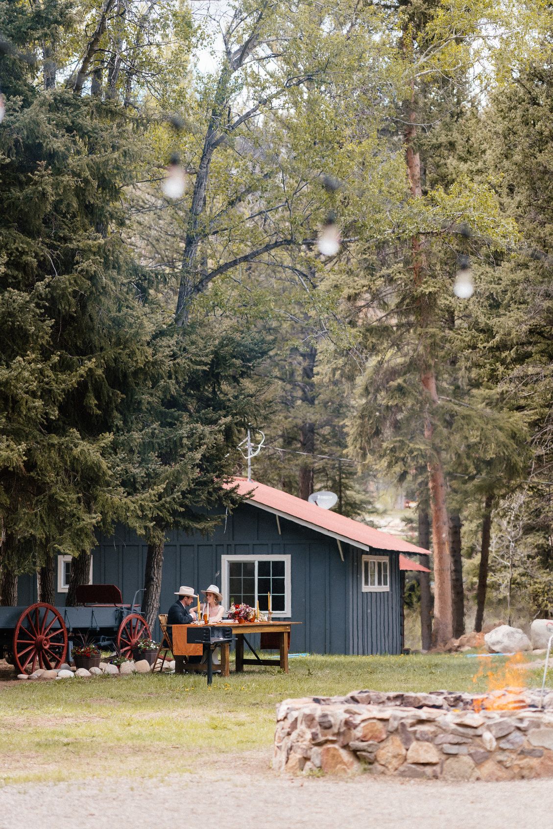 Guests at a table in front of a cabin, with the fire pit burning at the Cookout Grounds